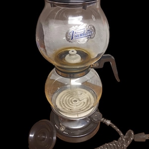 May include: A vintage Vaculator coffee maker with a clear glass carafe and a black base. The glass carafe has a black lid and a handle. The base has a switch labelled "Warm" and "Brew". The power cord is black and grey.