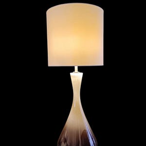 Puede incluir: Una lámpara de mesa de cerámica marrón y crema con una pantalla de tela blanca. La lámpara tiene una base ancha y un cuello estrecho.