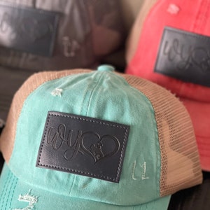 May include: Three trucker hats with distressed denim and mesh panels. The hats are in different colors: gray, teal, and coral. Each hat has a black leather patch with the word "why" and a heart design.