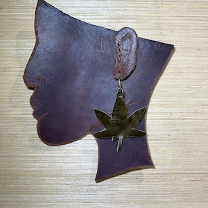 May include: A brown leather silhouette of a person's head with a gold leaf earring. The leaf is a stylized marijuana leaf.