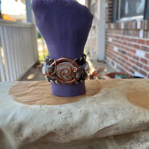 Brazalete de cuero marrón con banda ancha, asegurado con cobre desnudo y cuero, conchas cauri, mezclilla clara y encuadernado en espiral de cáñamo con broches personalizados.