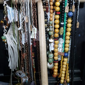 May include: A variety of necklaces and chains hanging on a wooden rack. The necklaces are made of different materials, including beads, stones, and metal. Some of the necklaces have tags with prices on them.