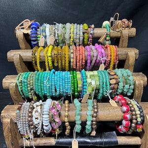 May include: A wooden display stand with multiple rows of colorful beaded bracelets. The bracelets are made of various materials, including glass, wood, and metal. Some of the bracelets have price tags attached.