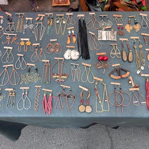 May include: A collection of various handmade earrings displayed on small rectangular cardboard cards. The assortment includes wire-formed shapes, long fringe tassels in red and black, geometric metal designs, and natural elements like wood and beads.