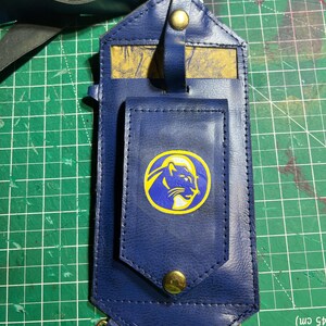 Puede incluir: Una funda de teléfono de cuero azul marino con un cierre dorado y un emblema de león dorado sobre un círculo azul y amarillo. La funda tiene una correa y un clip de metal dorado.