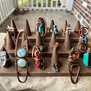 May include: A display of handcrafted rings with various gemstones and wire-wrapped designs. The rings are copper-toned and feature stones in shades of blue, green, red, and clear. They are arranged on a wooden display stand with brown, cone-shaped ring holders.