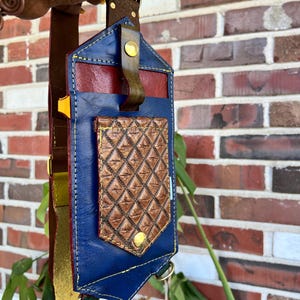 Könnte beinhalten: Handgefertigte Leder-Umhängetasche in Blau, Rot und Braun. Sie hat eine diamantgemusterte Vordertasche und goldfarbene Hardware. Der Riemen kombiniert Braun, Gelb und Gold. Ein stilvolles Accessoire.