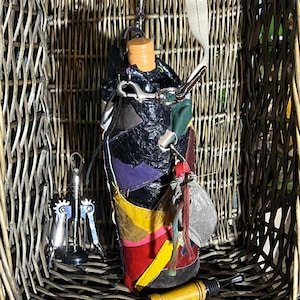 May include: A colourful, patchwork wine bottle with a cork stopper and a leather strap. The bottle is in a wicker basket with a corkscrew.