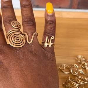 May include: A collection of gold-toned wire rings featuring various designs, including a large spiral, a V-shape, and a letter N. Several additional wire rings with similar spiral and abstract shapes are arranged in a small pile nearby.