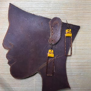 May include: A pair of handmade earrings with yellow accents and copper-colored rectangular frames. The earrings are displayed on a brown leather silhouette of a face and ear. The background is a light wood grain.