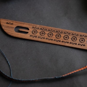 Peut inclure: Un outil de tissage en bois avec un motif sculpté de cercles et de volutes. L'outil comporte deux trous pour enfiler le fil.