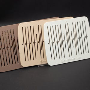May include: Three square, slotted wooden panels in shades of brown, beige, and white. Each panel has vertical slots and small holes. The white panel has the text "@nicoypoodyak". A patterned strap is visible in the background.