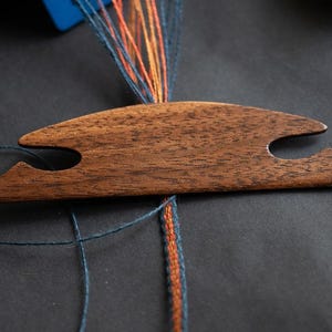 Può includere: Uno strumento in legno con due fori per tessere fili. Lo strumento è marrone e ha una superficie liscia. È posto su una superficie grigio scuro con fili blu, arancioni e rossi.