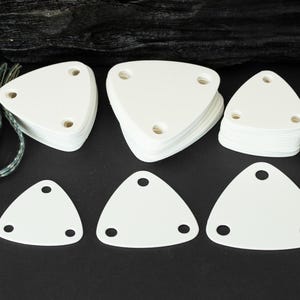 May include: White triangular tags with three holes, arranged on a black surface. Several stacks of tags are visible, along with individual tags. A spool of thread and a length of braided cord are also present.