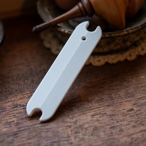 May include: A white plastic sewing gauge with a hole in the centre and a series of lines running down the length of the gauge.