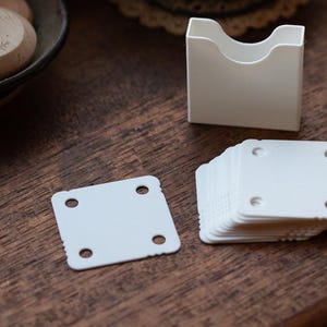 May include: A white business card holder and a stack of white square cards with rounded corners and four holes. The cards are on a dark wooden surface. A wooden bowl with wooden objects is in the background.