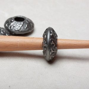 Puede incluir: Un huso de rueda de hilar de metal plateado con un dise&ntilde;o floral decorativo. El huso est&aacute; en un husillo de madera.