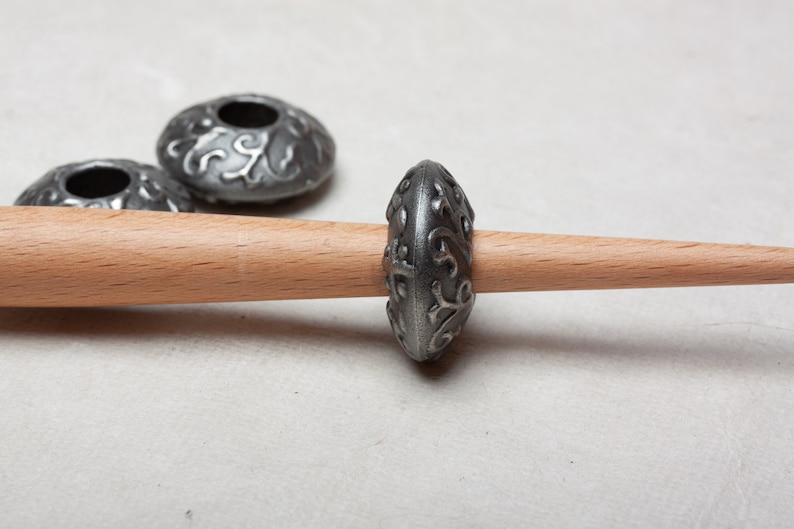Puede incluir: Un huso de rueda de hilar de metal plateado con un dise&ntilde;o floral decorativo. El huso est&aacute; en un husillo de madera.
