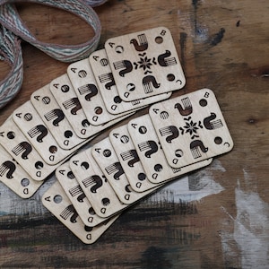 May include: A stack of wooden knitting stitch markers with a bird and flower design. The markers are labelled with the letters A, B, C, and D.