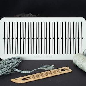 May include: A white weaving loom with vertical slots and the text "funkyneedleduk". Accessories include a woven band, a wooden shuttle, and two spools of yarn in grey and teal. The items are arranged on a black surface.