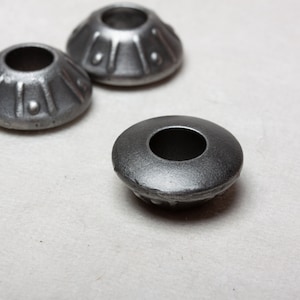 Op de afbeelding: Drie zilveren metalen meubellegdoppen met een decoratief ontwerp. De doppen hebben een ronde bovenkant met een gat in het midden en een geribbelde basis.