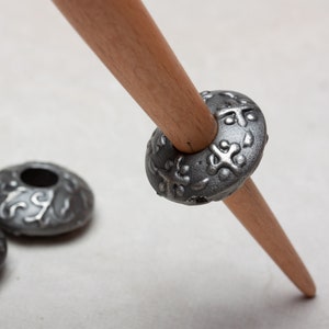 Puede incluir: Un huso de hilar de madera con tres hileras de metal plateado. Las hileras tienen un dise&ntilde;o decorativo.
