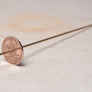 Może przedstawiać: Moneta miedziana z wzorem ptaka i tekstem "1p" oraz "ONE PENNY" przymocowana do cienkiego, złotego pręta metalowego.