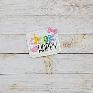 May include: A white paper clip with a white square attached to it. The square has a pink bow and the words "choose happy" in rainbow colors. The words are embroidered on the square.