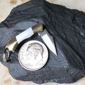 May include: A small, white and gold pocket knife with a mother-of-pearl handle. The knife is open and the blade is visible. The knife is next to a US dime coin for scale.