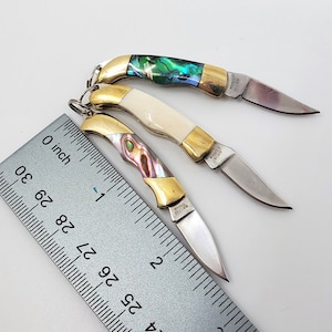 May include: Three small pocket knives with different colored handles. The handles are made of abalone shell and have gold accents. The blades are all closed and measure about 2 inches long. The knives are laid out on a white surface next to a ruler.