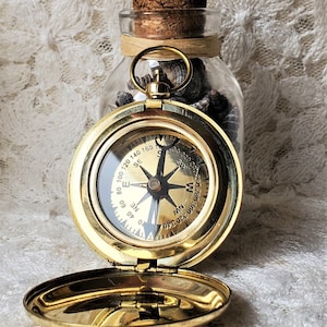 May include: A gold compass with a glass face and a black needle. The compass is open and the lid is laying next to it. The compass is sitting on a white lace surface. The compass has the numbers 0-360 around the edge and the letters N, E, S, and W in the center.