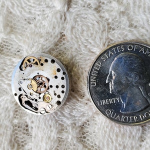 Pu&ograve; includere: Un movimento di orologio smontato con ingranaggi e componenti intricati, accanto a un quarto di dollaro americano per il confronto delle dimensioni. Il meccanismo dell'orologio &egrave; argentato e dorato, mostrando i delicati meccanismi interni di un orologio. La moneta mostra il profilo di George Washington.