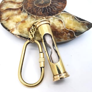 May include: A brass hourglass keychain with a white sand timer inside. The keychain is attached to a brass key ring.