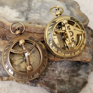 May include: Two antique brass pocket sundials with compass features. The sundials have Roman numeral markings and a compass rose with directional markings. The sundials are on a brown and white stone surface.