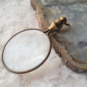 May include: A brass magnifying glass with a round lens and a decorative handle. The magnifying glass is on a white surface.