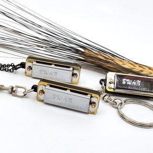 May include: Two silver and gold harmonicas with the text "SWAN HARMONICA" on them. One harmonica is attached to a silver chain and the other is attached to a silver key chain.