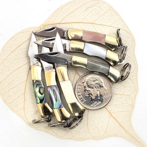 Puede incluir: Cinco pequeñas navajas de bolsillo con mangos dorados e incrustaciones de diferentes colores. Las navajas están apiladas y tienen llaveros. Se incluye una moneda de diez centavos de dólar estadounidense para la escala.