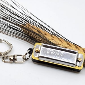 May include: A silver and gold harmonica keychain with the word "SWAN" engraved on it. The keychain is attached to a wheat stalk with a silver key ring.