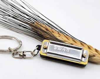 May include: A silver and gold harmonica keychain with the word "SWAN" engraved on it. The keychain is attached to a wheat stalk with a silver key ring.
