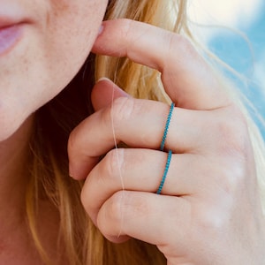 Op de afbeelding: Twee dunne gouden ringen met een rij turquoise blauwe stenen. De ringen worden gedragen aan de ringvinger van een hand.