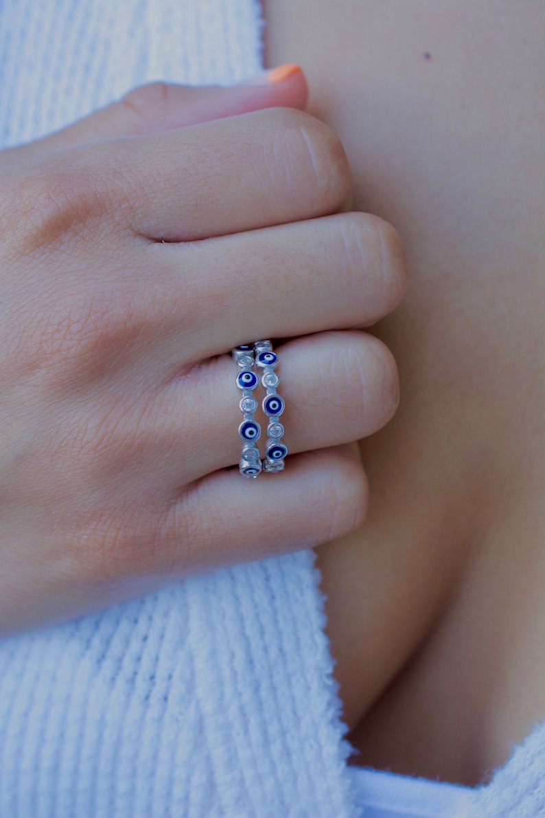 May include: A silver ring with a double row of blue evil eye charms. Each charm has a white center and is surrounded by small clear stones.