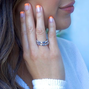 May include: A silver ring with a double row of blue evil eye charms. The ring is worn on the index finger of a person with orange nail polish.