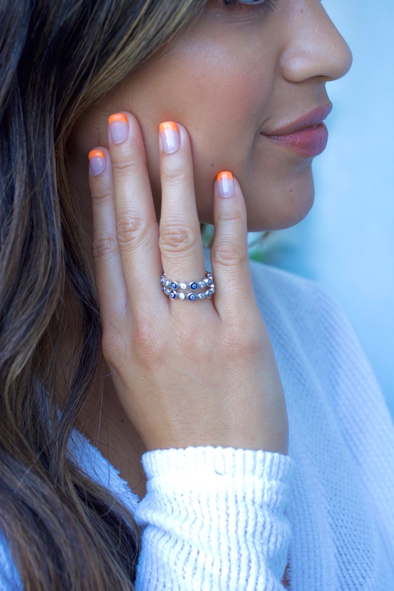 May include: A silver ring with a double row of blue evil eye charms. The ring is worn on the index finger of a person with orange nail polish.