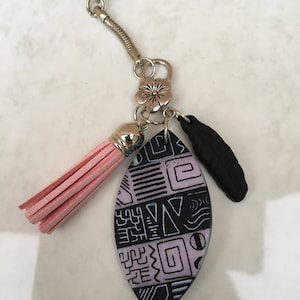 Puede incluir: Un llavero ovalado con estampado negro y morado con un borla rosa, un dije de pluma negra y un dije de espiral plateado. El llavero está unido a un anillo de resorte plateado y un anillo de llave plateado.