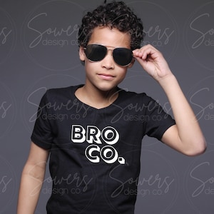 Może przedstawiać: Młoda osoba w czarnym t-shircie z napisem "BRO CO." w kolorze białym. Osoba nosi okulary przeciwsłoneczne i ma krótkie kręcone włosy.