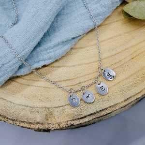 May include: Silver necklace with four charms: a heart, a letter D, a letter E, and a leaf. The necklace is on a wooden surface.