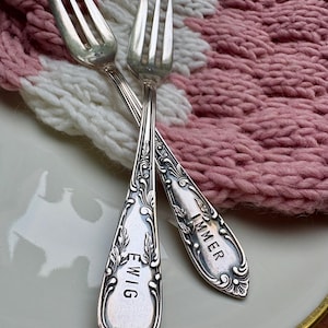 May include: Two silver forks with decorative handles, one inscribed with "EWIG" and the other "IMMER". The forks are crossed on a white plate, with a pink and white knitted textile in the background. The forks have a vintage appearance.