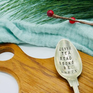Könnte beinhalten: Ein silberner Löffel mit der Aufschrift "DRINK TEA READ BOOKS BE HAPPY" liegt auf einem Holzschneidebrett. Eine blumige Teetasse und ein Zweig Grünzeug sind im Hintergrund. Ein charmantes Küchenutensil.