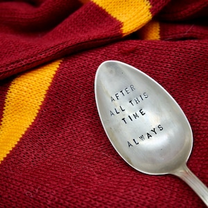 Könnte beinhalten: Ein silberner Löffel mit dem eingestanzten Schriftzug "AFTER ALL THIS TIME ALWAYS". Der Löffel liegt auf einem rot-gold gestreiften Strickstoff, was eine warme, thematische Präsentation ergibt.