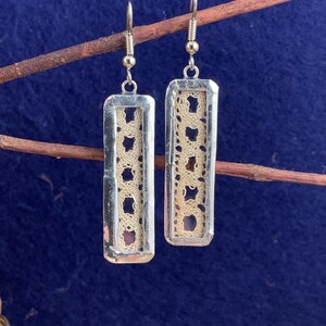 May include: Silver-toned rectangular earrings with a cream-colored lace insert. The earrings hang from silver hooks and are displayed against a blue background. The lace has a repeating pattern of small, open shapes.